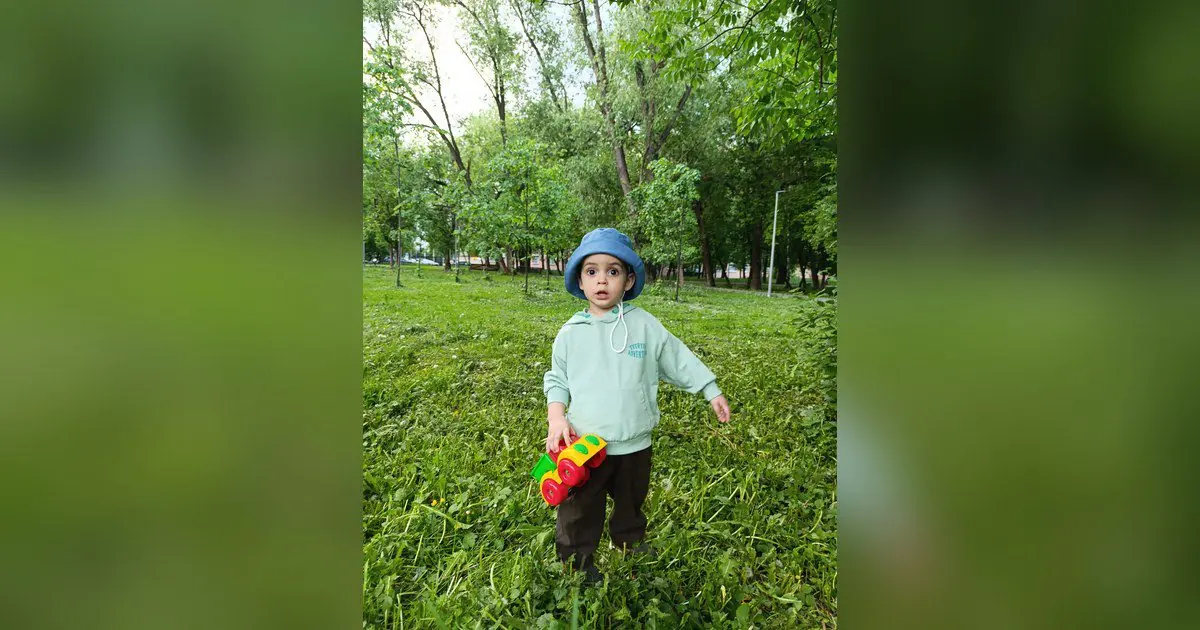 Ilie, băiat de 2 ani, cu pălărie bleu și un trenuleț de jucărie, în parc